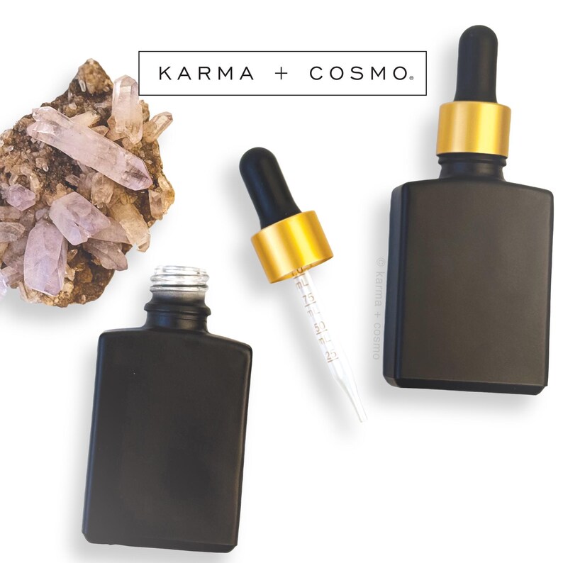 May include: Black square essential oil bottles with gold and black droppers. The bottles are displayed with a cluster of quartz crystals and the text "KARMA + COSMO."