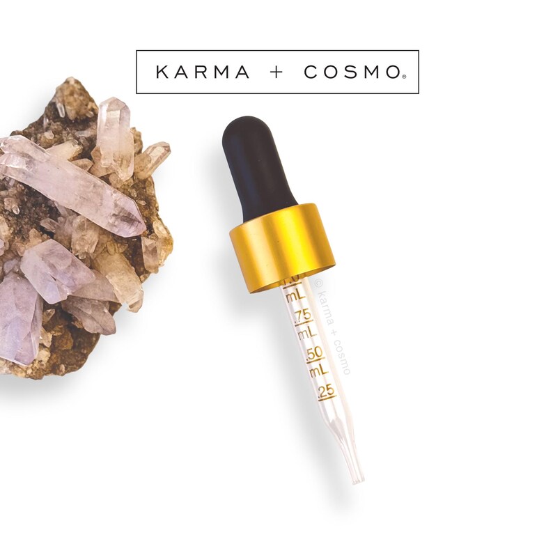 May include: A clear glass dropper with a black top and gold collar, marked with mL measurements. The dropper is angled against a white background, with a cluster of crystals and the text "KARMA + COSMO" at the top.