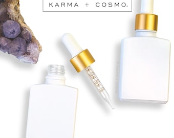 May include: White square essential oil bottles with gold accents and droppers. The bottles are displayed with a crystal and the text "KARMA + COSMO."