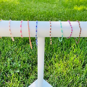 May include: A white display stand with eight colorful beaded bracelets. The bracelets are made of small, colorful beads and are arranged in a row on the stand.