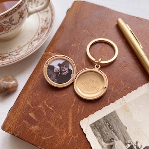 May include: A gold-toned metal locket keyring featuring a circular frame with a hinged opening. The left side contains a black and white photograph of two people, while the right side is engraved with the text always with you in a serif font.
