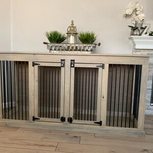 May include: A double-door dog crate made of wood with a light gray stain and black metal bars. The crate has two doors with black metal handles and hinges.