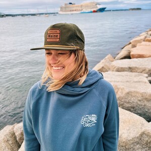 May include: A woman wearing a blue hooded sweatshirt with a white "East Coast" logo and a green baseball cap with a brown "State Line Maine" patch. She is standing on a rocky shore with a cruise ship in the background.