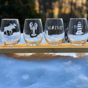 May include: Four clear glass wine glasses with etched designs. The glasses feature a moose, a lobster, the state of Maine, and a lighthouse.