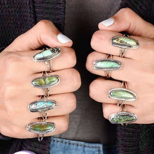 May include: A hand model wearing multiple silver rings with turquoise stones. The rings are stacked on each finger and feature different shapes and sizes of turquoise.