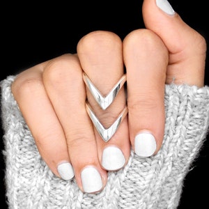 May include: A silver double chevron ring with a minimalist design. The ring is worn on the index finger of a hand with white nail polish.