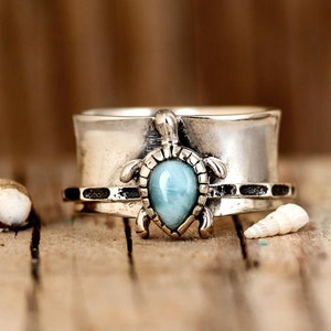 May include: A silver ring with a blue gemstone set in a turtle design. The ring is wide and has a textured band.