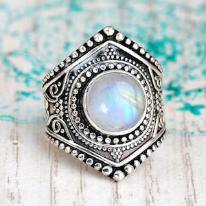 Pode incluir: Um anel de prata com uma grande pedra de lua redonda e iridescente engastada em um design detalhado e ornamentado.