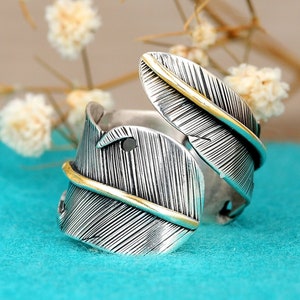 May include: A silver ring with a gold feather design. The ring is open and adjustable.