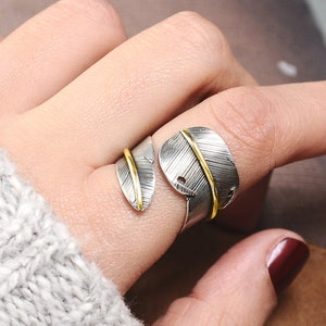 May include: A silver ring with a gold accent, shaped like two overlapping leaves. The ring is worn on a finger.