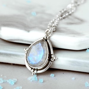 May include: A silver pendant necklace featuring a pear-shaped, iridescent gemstone. The gemstone is set in a silver bezel with decorative accents. The necklace chain is delicate and silver. The overall design is elegant and suitable for everyday wear.