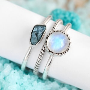 May include: Three silver rings with gemstones. The ring on the left has a blue gemstone. The middle ring has a white gemstone with a silver setting. The ring on the right is a plain silver band.
