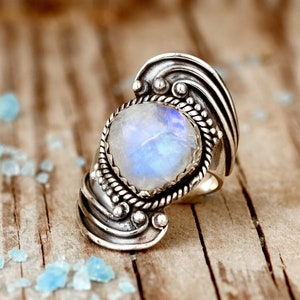 May include: A silver ring with a large, oval moonstone. The moonstone has a pearly white and blue iridescence, set in a detailed, ornate silver band with a textured design. The ring is on a wooden surface.