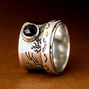 May include: A silver ring featuring a black gemstone and a textured design. The ring is crafted from two bands, one with a tree design and the other with a smooth surface.