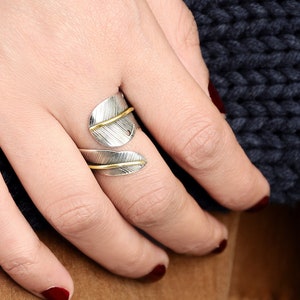May include: A silver and gold ring with a leaf design. The ring is worn on the index finger of a hand.