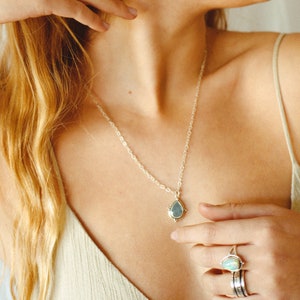 May include: A silver necklace with a teardrop-shaped pendant featuring a light blue stone. A silver ring with a white stone is also visible. The jewelry is worn against a person's skin, with a neutral-colored top.
