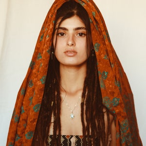 May include: A woman wearing a rust-colored headscarf with teal floral patterns. She has long, dark dreadlocks and is wearing a silver necklace with a teardrop-shaped pendant. The top of her dress is visible, featuring an intricate black and gold design.