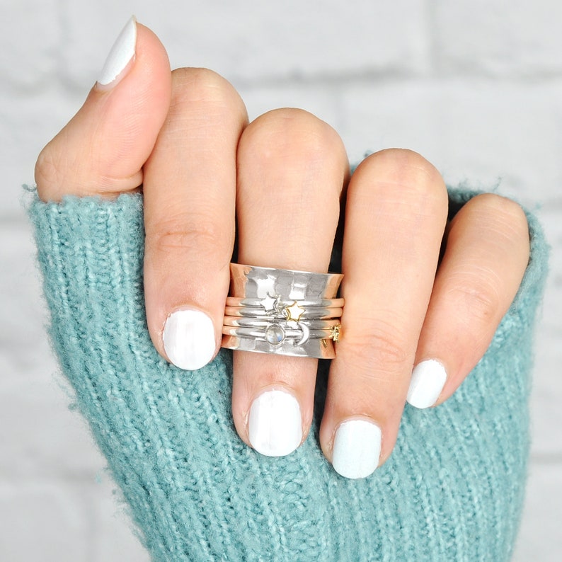 May include: A hand wearing a stack of silver and gold rings with a star and moon design. The rings are on the index finger and are worn on top of a turquoise blue knitted sweater.