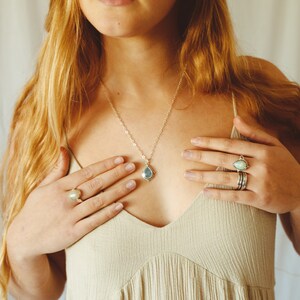 May include: A silver necklace with a teardrop-shaped pendant and two rings with light blue gemstones. The rings are different styles, one with a single stone and the other with multiple bands. The jewelry is displayed on a person wearing a beige dress.