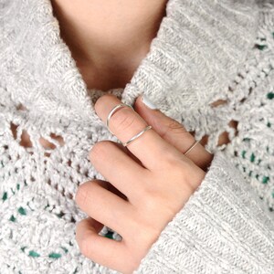 May include: A hand wearing three silver rings. One ring curves over the top of the finger, another is a simple band, and the third is a thin, twisted band. The person is wearing a gray knit sweater.