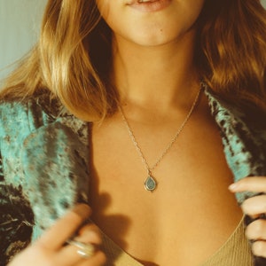 May include: A gold-colored necklace with a teardrop-shaped pendant. The pendant has a light blue center and a decorative border. The necklace is worn against a person's skin, with a teal and black patterned jacket in the background.