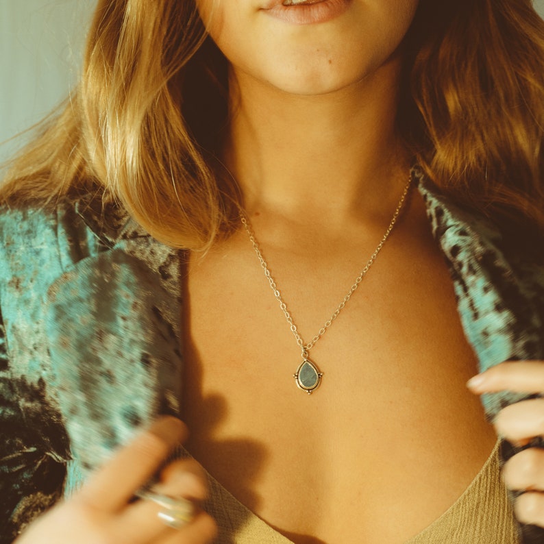 May include: A gold-colored necklace with a teardrop-shaped pendant. The pendant has a light blue center and a decorative border. The necklace is worn against a person's skin, with a teal and black patterned jacket in the background.