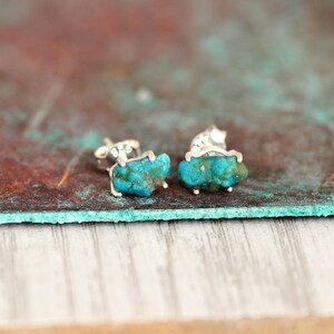 May include: A pair of silver earrings featuring turquoise stones. The stones are a deep blue-green colour and have a rough, natural texture.