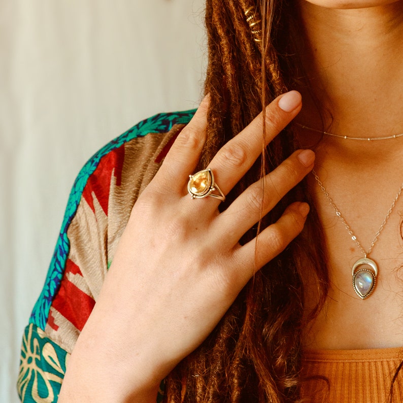 May include: A teardrop-shaped ring with a yellow gemstone set in a silver band. The ring is worn on the index finger of a hand with long brown dreadlocks.