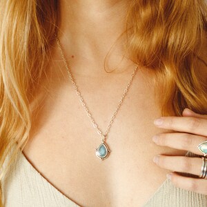 May include: A silver necklace with a teardrop-shaped pendant. The pendant features a light blue gemstone with a silver border and small decorative accents. The necklace is worn around the neck.