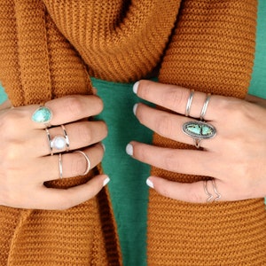 May include: A collection of silver rings with turquoise and moonstone accents. The rings are worn on multiple fingers, showcasing various designs, including a teardrop-shaped turquoise ring and a ring with a large oval turquoise stone.