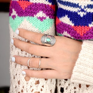 May include: A hand wearing two rings. One is a wide, silver ring with a turquoise stone and geometric designs. The other is a thin, silver band. The person is wearing a colorful, patterned sweater with white nail polish.