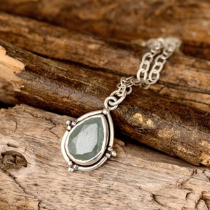 May include: A silver necklace with a teardrop-shaped pendant. The pendant has a light green gemstone set in a silver frame with decorative accents. The necklace chain is also silver and rests on a wooden surface.