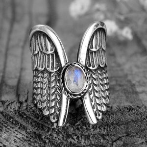 May include: A silver ring with a large, round, iridescent gemstone set in the center. The ring is shaped like a pair of wings.