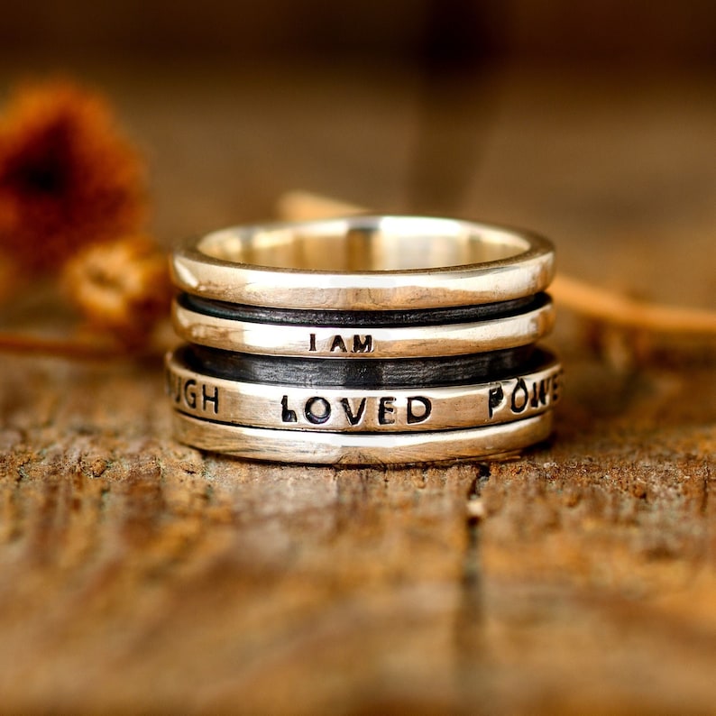 May include: A silver ring with four bands, each engraved with a different word. The words are "I AM", "LOVED", "POWER", and "ENOUGH".