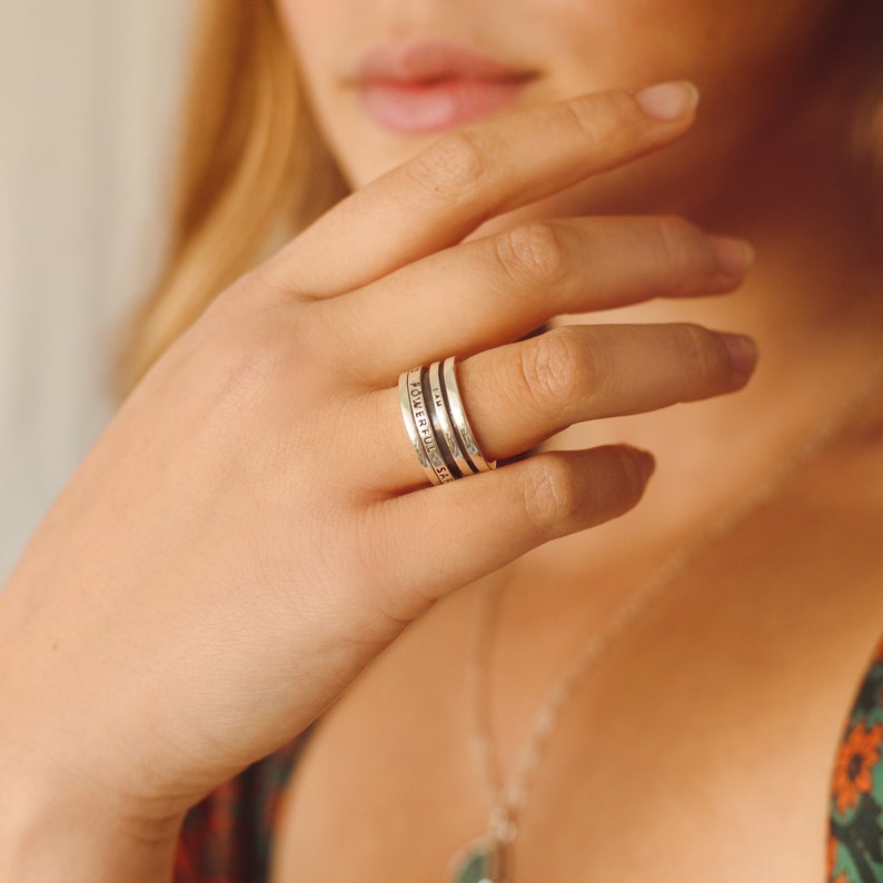 May include: A silver ring with the words "powerful" and "beautiful" engraved on it. The ring is worn on the middle finger of a person's hand.