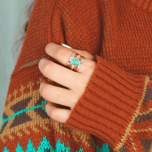 May include: A close-up of a hand wearing a silver ring with a turquoise stone. The ring is stacked with other silver bands. The hand is wearing a red and brown sweater with a geometric pattern.