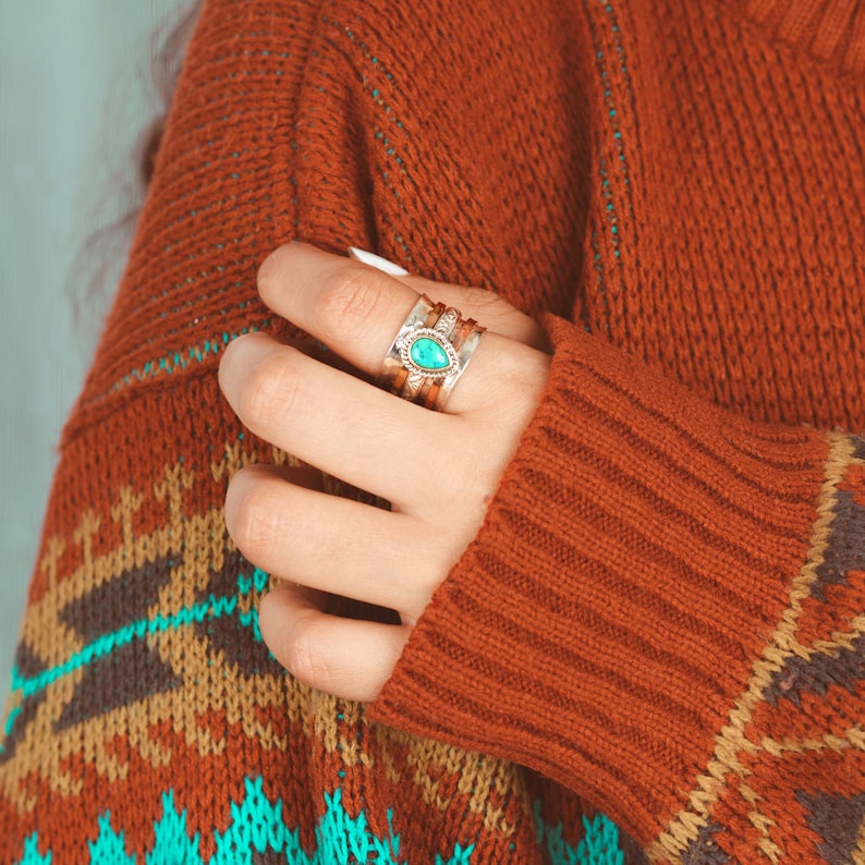 May include: A close-up of a hand wearing a silver ring with a turquoise stone. The ring is stacked with other silver bands. The hand is wearing a red and brown sweater with a geometric pattern.