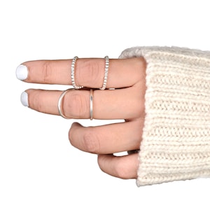 May include: A hand wearing four silver rings. Two rings are thin bands, and two are beaded. The hand is wearing a cream-colored knitted sweater. The fingernails are painted white.