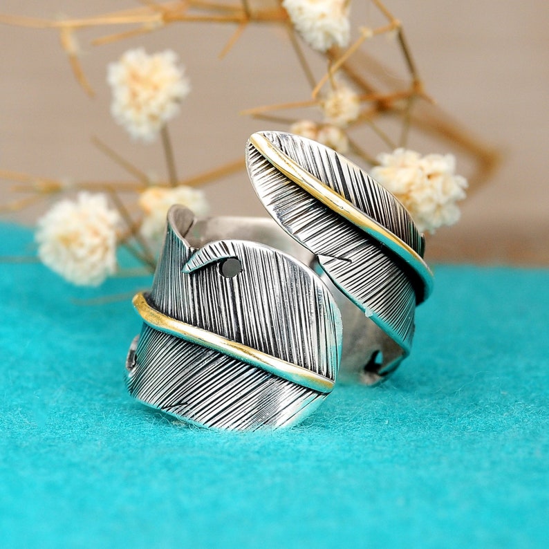 May include: A silver ring with a gold feather design. The ring is open and adjustable.
