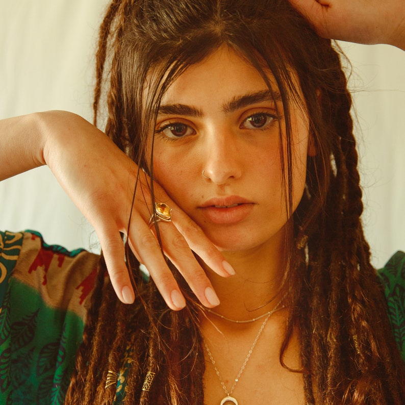 May include: A woman with long brown dreadlocks wears a green and brown patterned shirt. She has a gold ring with a yellow stone on her finger and a silver necklace with a circular pendant.