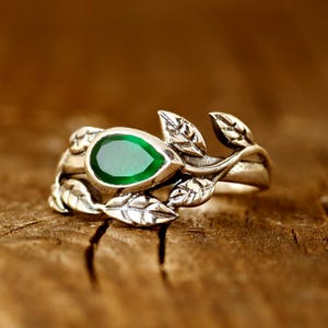 May include: A silver ring with a green gemstone and a leafy design. The ring is set on a wooden surface.