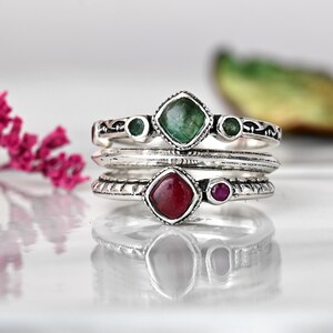 May include: A silver ring set with three green and two red gemstones. The ring is made of three bands, each with a different design. The largest gemstone is a square emerald, and the smaller gemstones are round rubies.