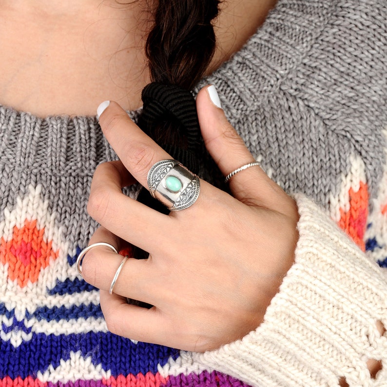 May include: A silver ring with an oval turquoise stone is worn on a finger. The ring has an ornate design with a rectangular turquoise stone in the center. A thin silver band ring and a twisted silver ring are also visible.