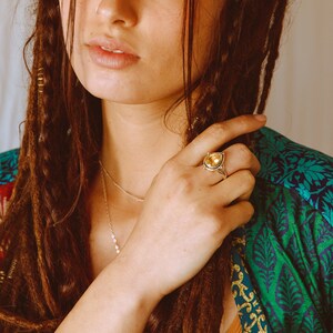May include: A woman wearing a green and gold patterned shirt with long brown hair. She is wearing a silver ring with a teardrop-shaped yellow gemstone.