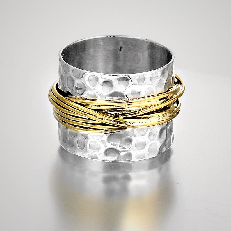 May include: A silver-colored ring with a hammered texture and a band of interwoven gold-colored wire. The ring has a wide band design and a polished finish, suitable for everyday wear.