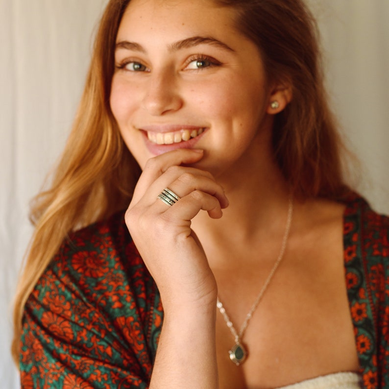 May include: A woman wearing a green and red patterned shirt with a silver necklace and a stack of silver rings on her finger.