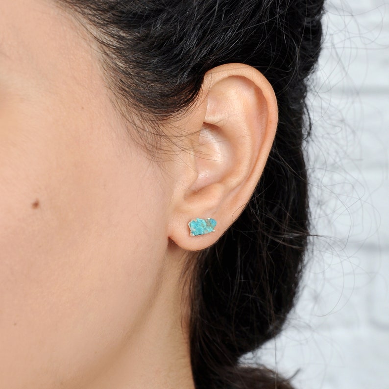 May include: A pair of turquoise stud earrings with a silver setting. The earrings are worn in a person's ear.