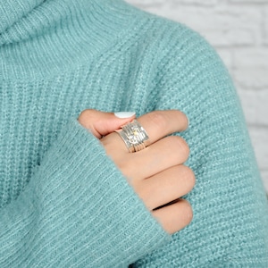 May include: A silver ring with multiple bands and small decorative accents. The ring is worn on a finger of a hand wearing a light blue sweater.