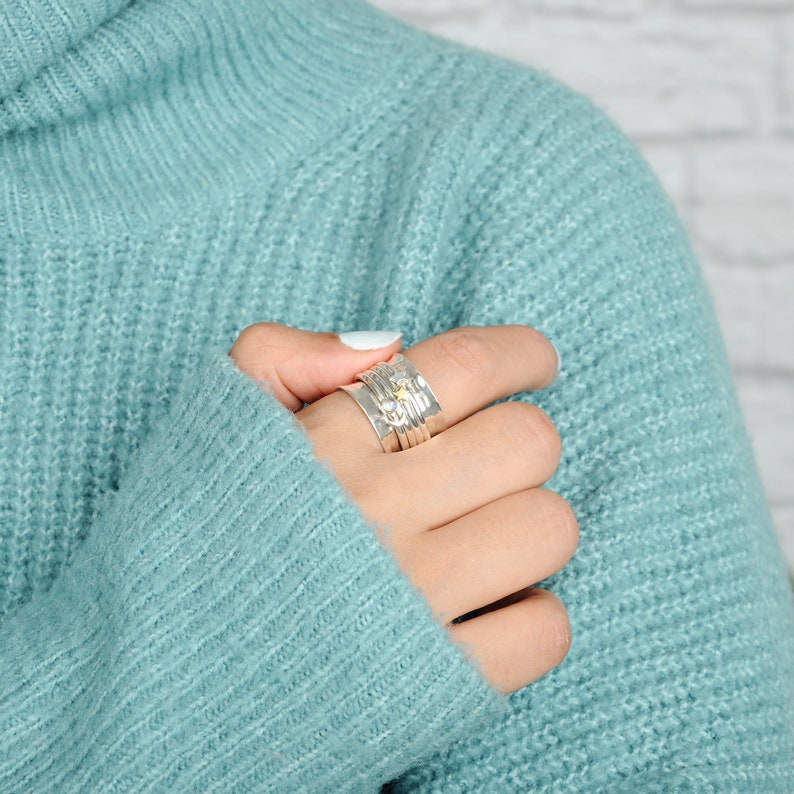 May include: A silver ring with multiple bands and small decorative accents. The ring is worn on a finger of a hand wearing a light blue sweater.