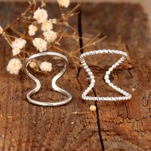 May include: Two silver rings are displayed on a wooden surface. One ring has a simple, curved design, while the other features a beaded pattern. The rings are set against a backdrop of dried flowers, creating a natural aesthetic.